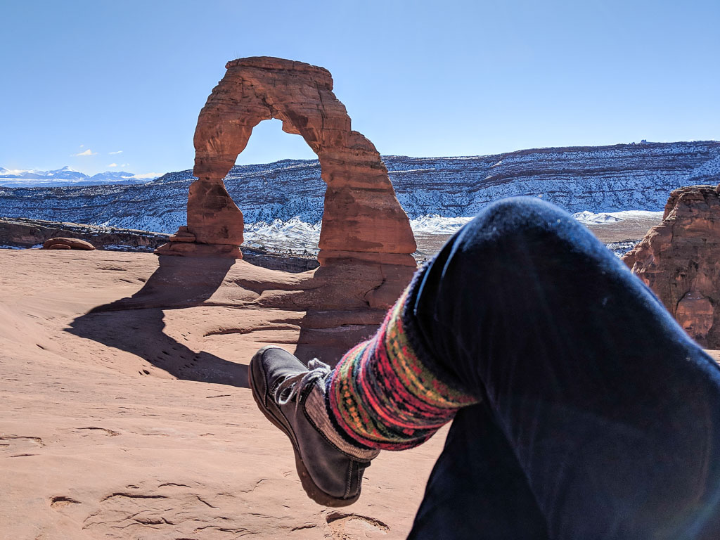 Molly's view of Delicate Arch at Arches National Park, with snow-covered mesa in the background and her leg in the foreground.